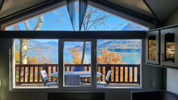 Habitación con ventanas amplias que ofrecen vistas a un lago y montañas. Hay una mesa y sillas afuera de las ventanas.