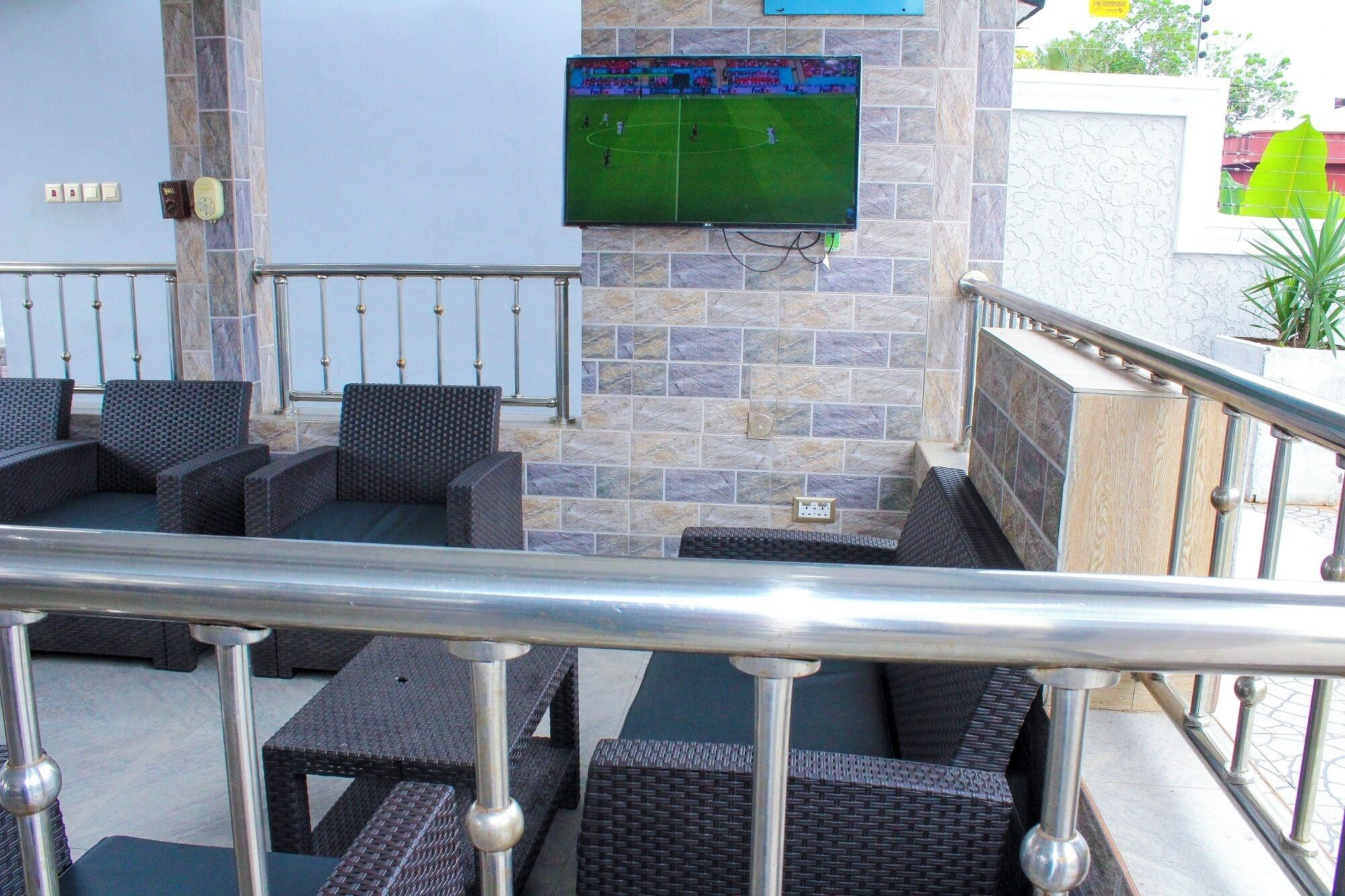 A terrace with wicker furniture, a television mounted on a stone wall, and a railing.