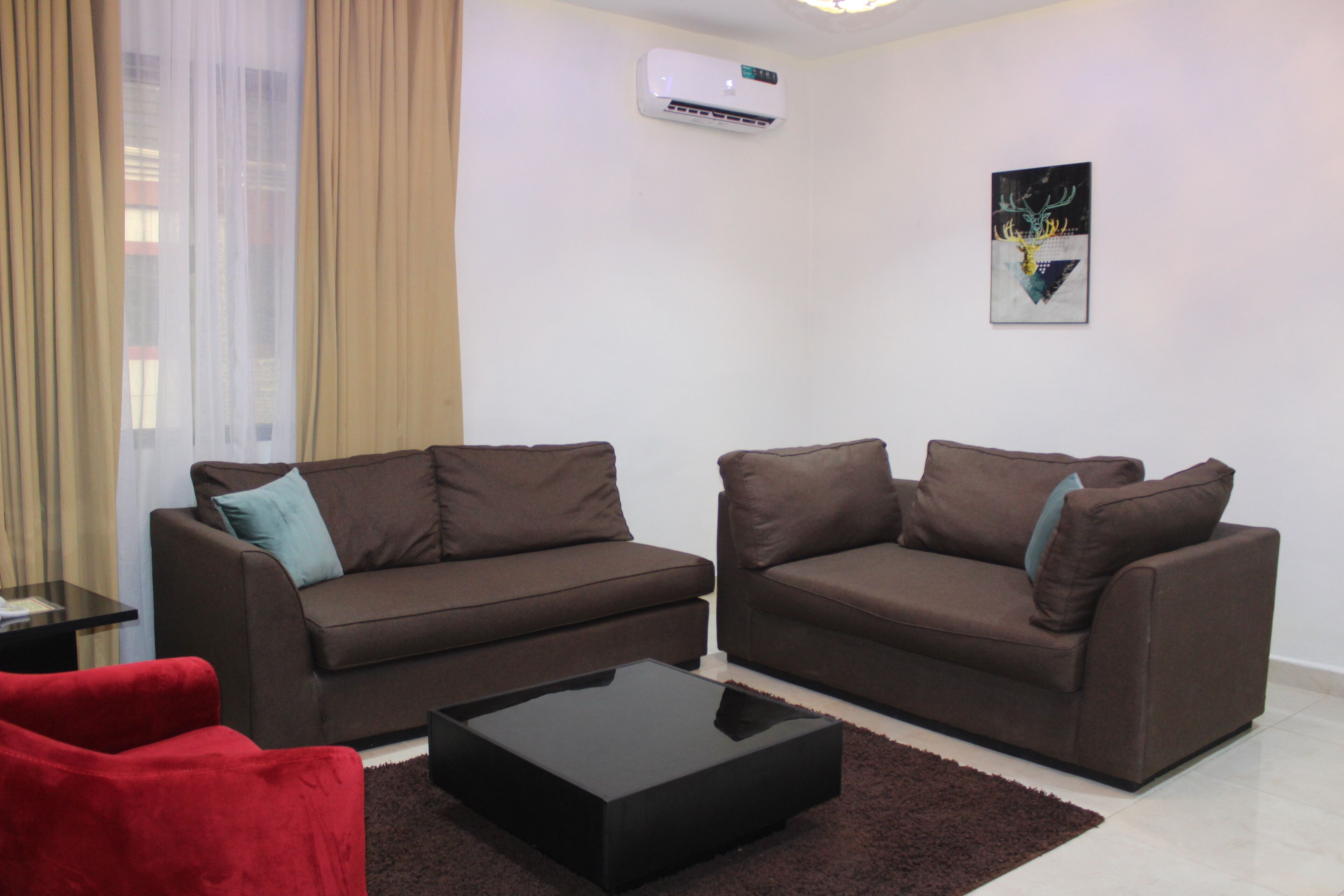 A living room with two brown sofas, a red chair, a black coffee table, a beige curtain, an air conditioner, and a wall art piece.