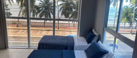 Habitación de hotel con dos camas, ventanas amplias y vistas a palmeras y la playa.