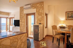 Ein Zimmer mit Steinsturz, einer kleinen Küchenzeile, einem Essbereich mit Stühlen, einem Fernseher und Meerblick durch eine Glastür.