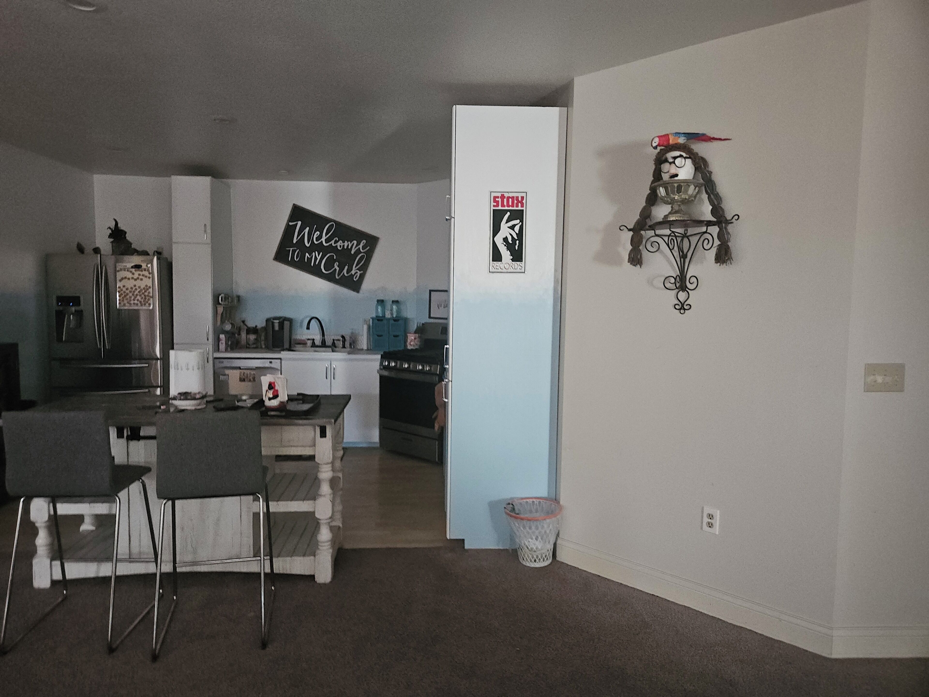 A modern kitchen with a refrigerator, stove, and sink. There is a dining area with chairs and a table. A sign on the wall reads "Welcome to My Crib."
