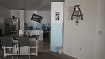 A modern kitchen with a refrigerator, stove, and sink. There is a dining area with chairs and a table. A sign on the wall reads "Welcome to My Crib."