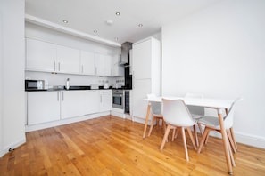 A modern kitchen with white cabinets, a black countertop, and a wooden floor. There is a dining table with white chairs. - Marylebone 2-Bed with Private Roof Terrace (London)