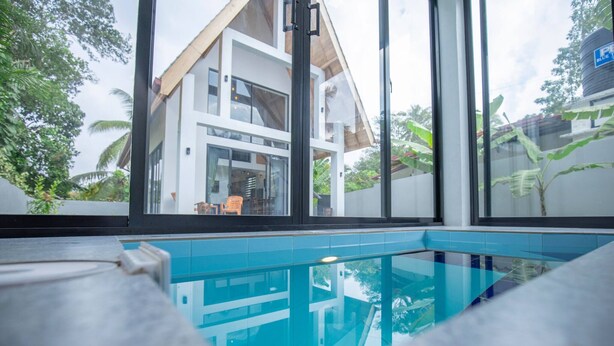 A modern pool area with clear glass walls, a view of a two-story house, and surrounding greenery.
