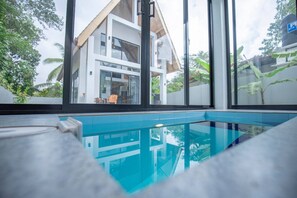 Area piscina moderna con pareti in vetro trasparente, vista su una casa a due piani e vegetazione circostante.