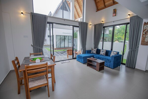 A modern living room with a dining area, large sliding glass doors leading to a patio, and a blue sofa with floral cushions. - Gone4coastal Cabana B (Hikkaduwa)