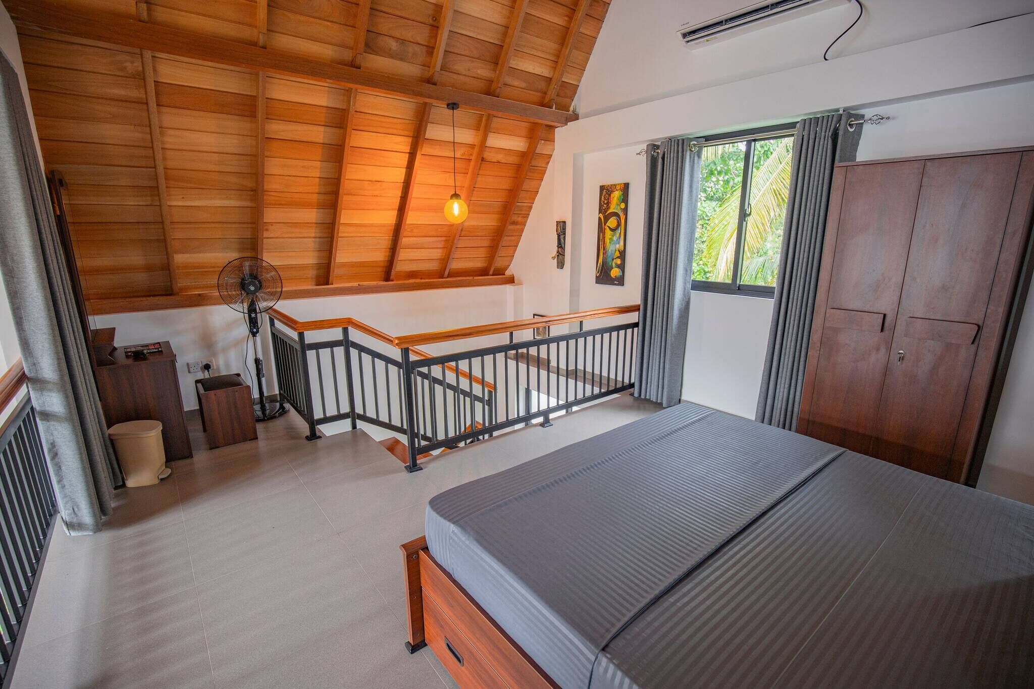 A bedroom with a wooden ceiling, a bed, a staircase, and a window with curtains.
