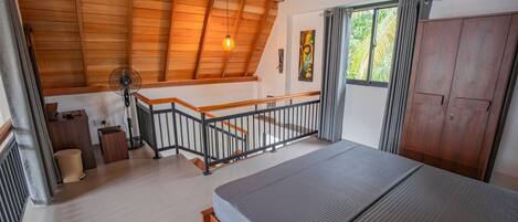 A bedroom with a wooden ceiling, a bed, a staircase, and a window with curtains.
