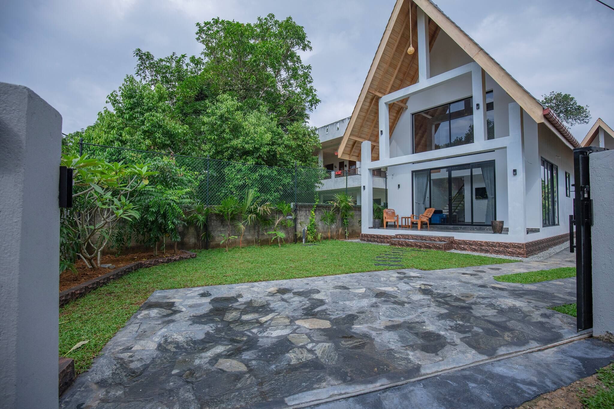 Una casa moderna de dos pisos con un patio de piedra, un jardín vallado y un árbol grande.