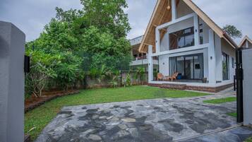 A modern two-story house with a stone patio, a fenced garden, and a large tree.