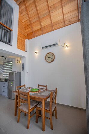 A dining area with a wooden table and chairs, a clock on the wall, and a kitchen in the background. - Gone4coastal Cabana B (Hikkaduwa)