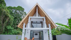 A modern two-story house with a wooden gable roof, a balcony with chairs, and a small TV screen.