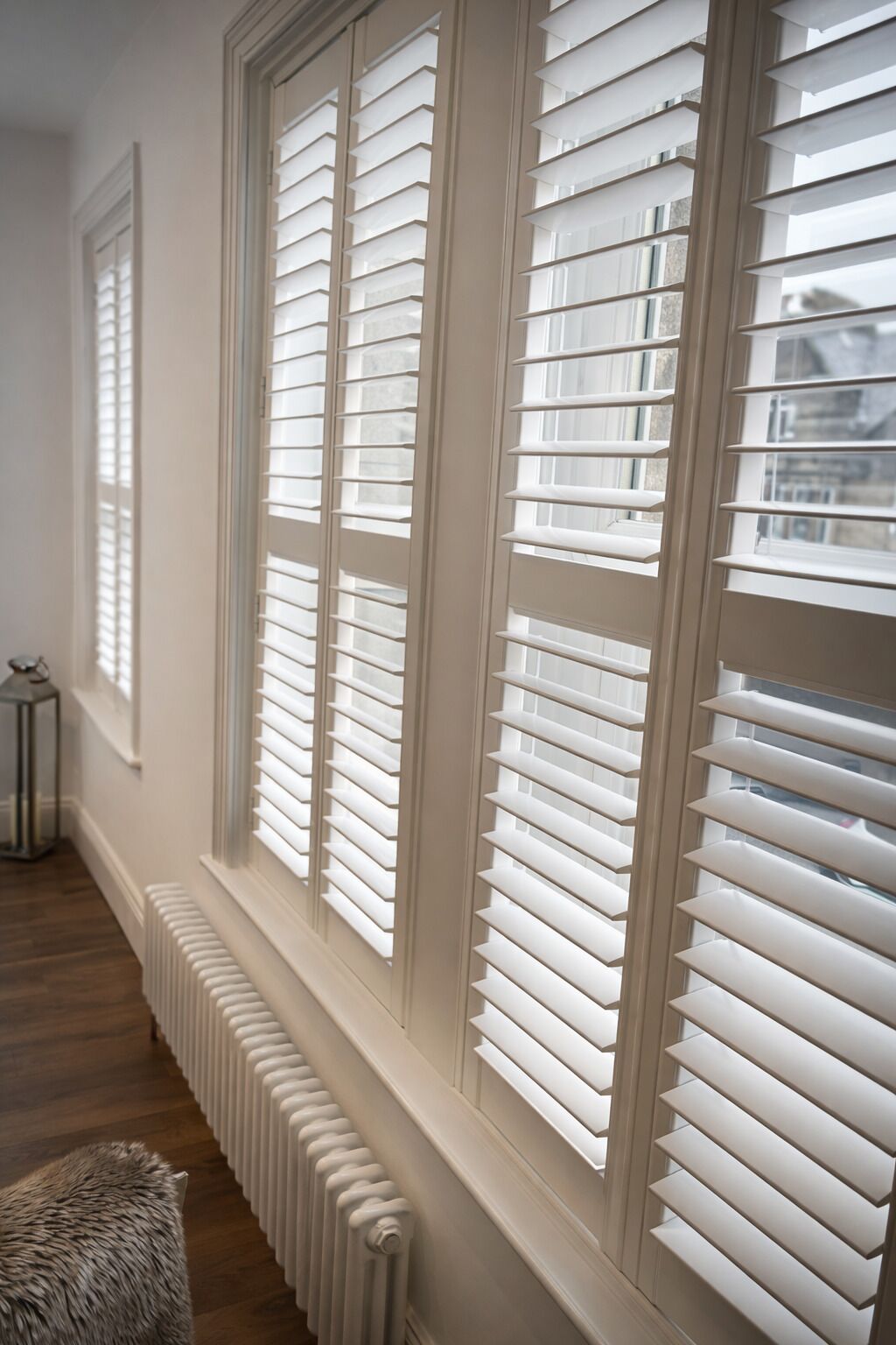 A hallway with large windows featuring white blinds, a radiator, and a fur-covered chair.