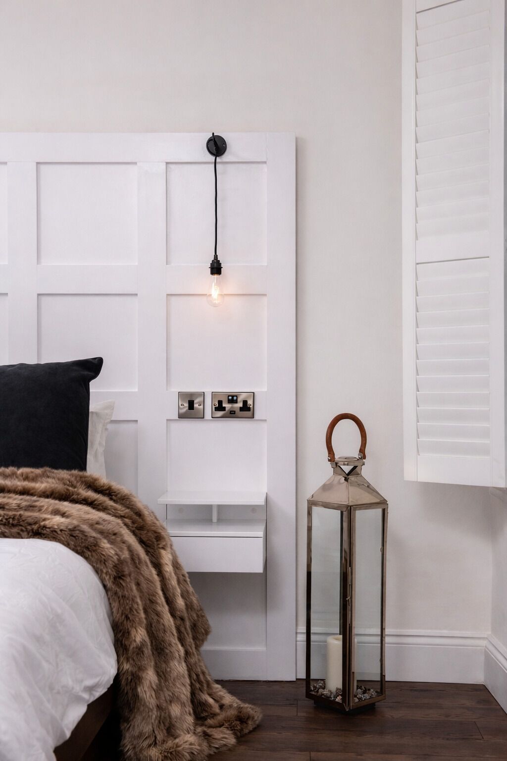 A bedroom with a white headboard, a bedside table with a lamp, a fur throw, and a lantern.
