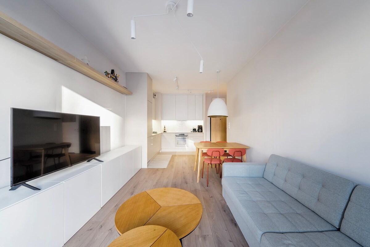 A modern living room with a flat-screen TV, a round wooden coffee table, a grey sofa, and a dining area with a table and chairs.
