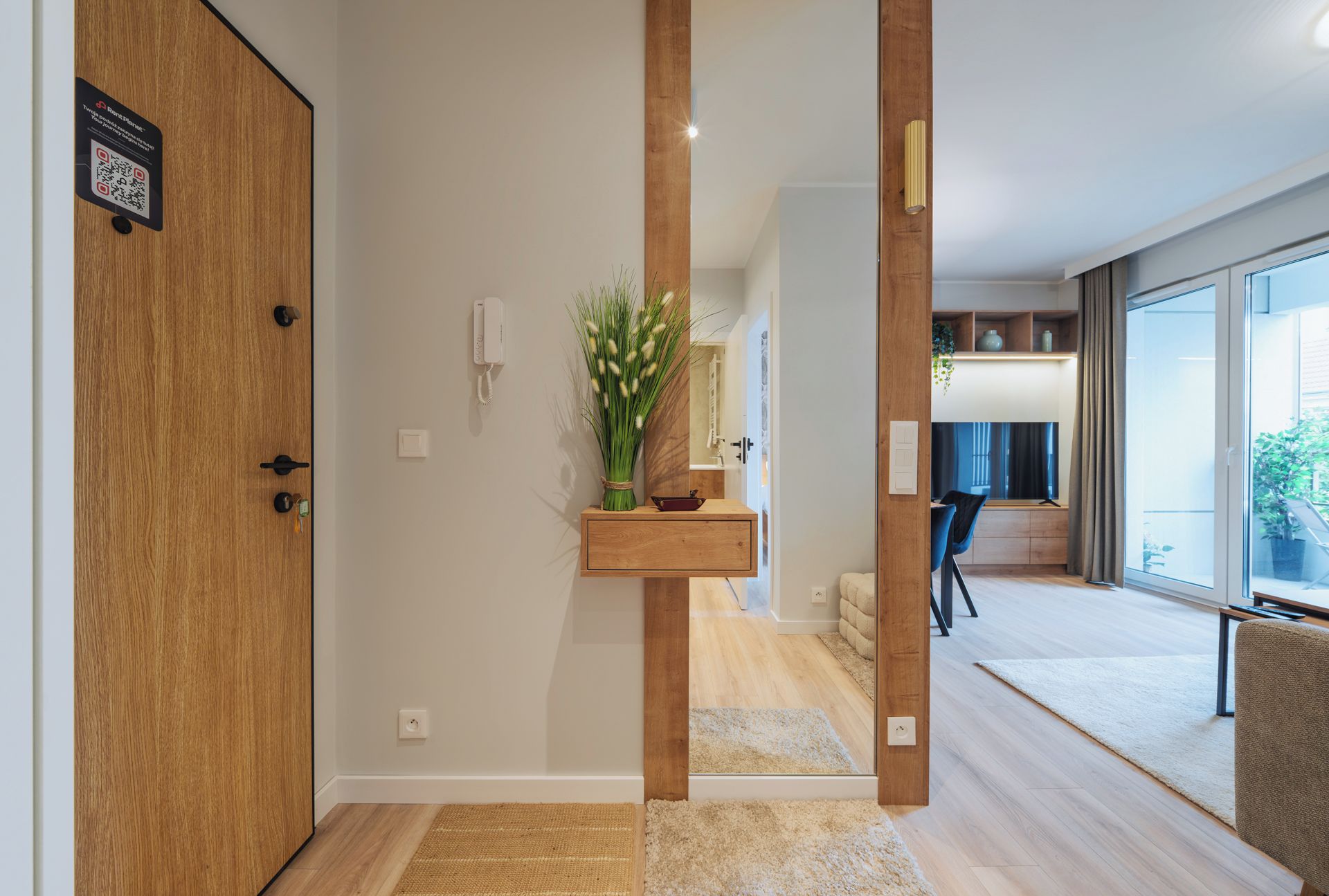 A hallway with a wooden door, a phone, a small wooden shelf with a plant, and a view into a room with a large window and a chair.