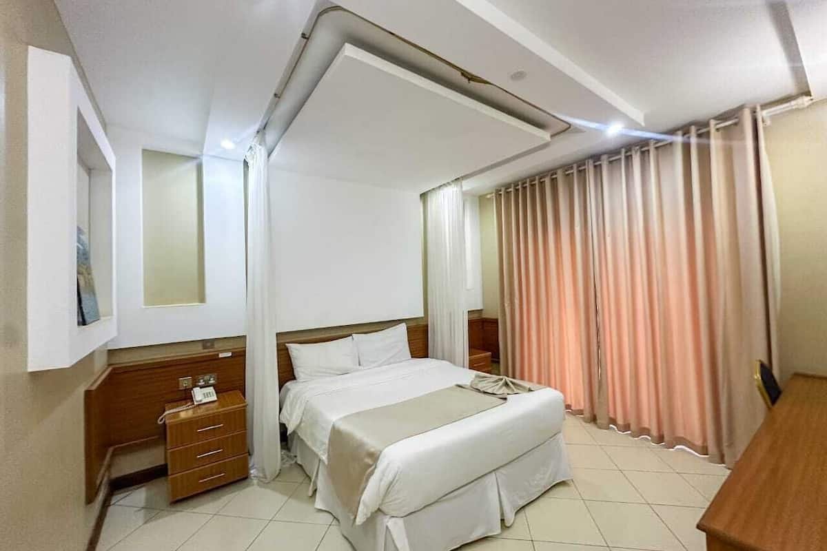 A hotel room with a bed, a wooden nightstand, a telephone, and a window with curtains.