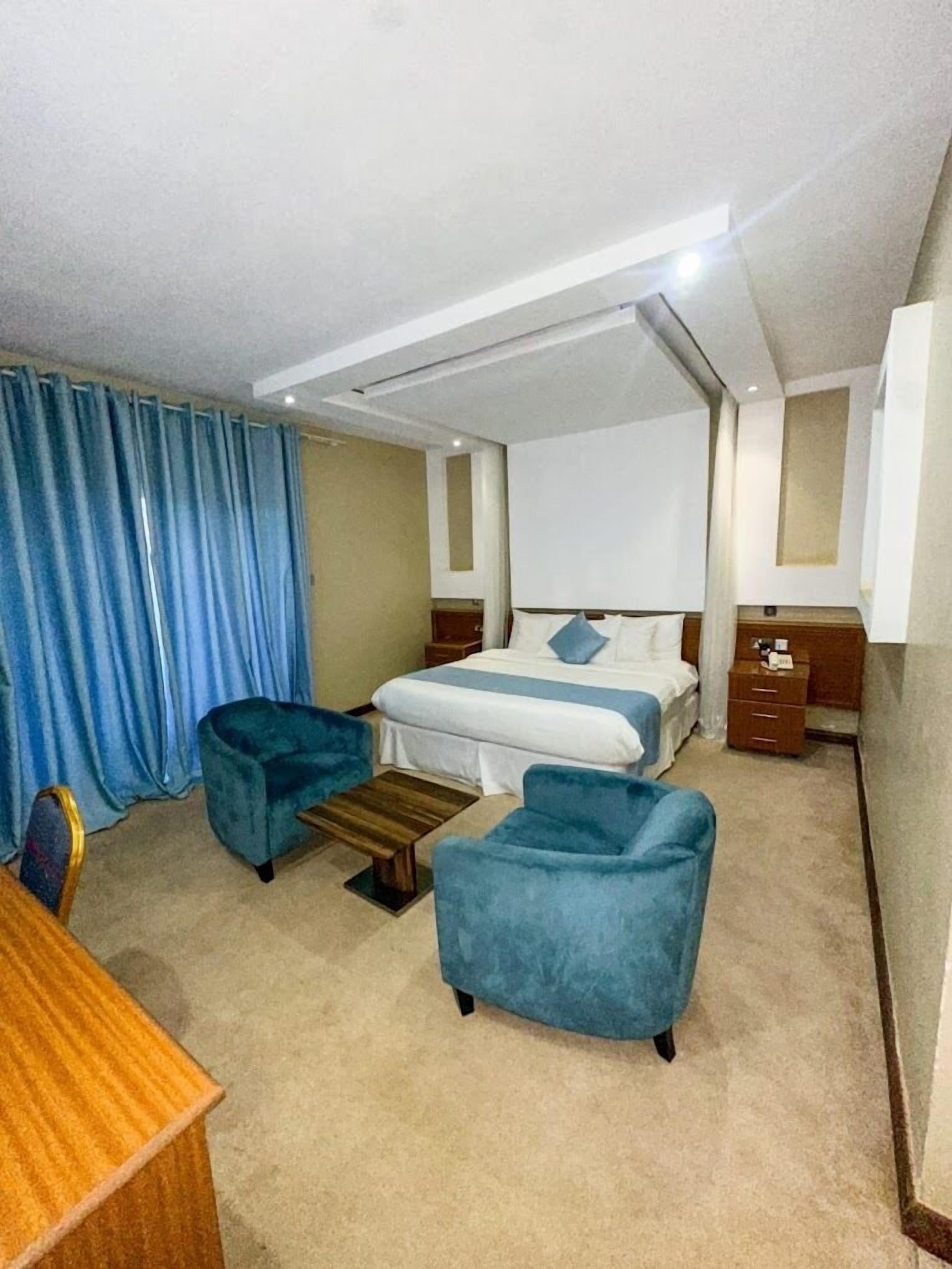 Ein Hotelzimmer mit einem Bett, zwei blauen Sesseln, einem Holztisch und einem kleinen Tisch.