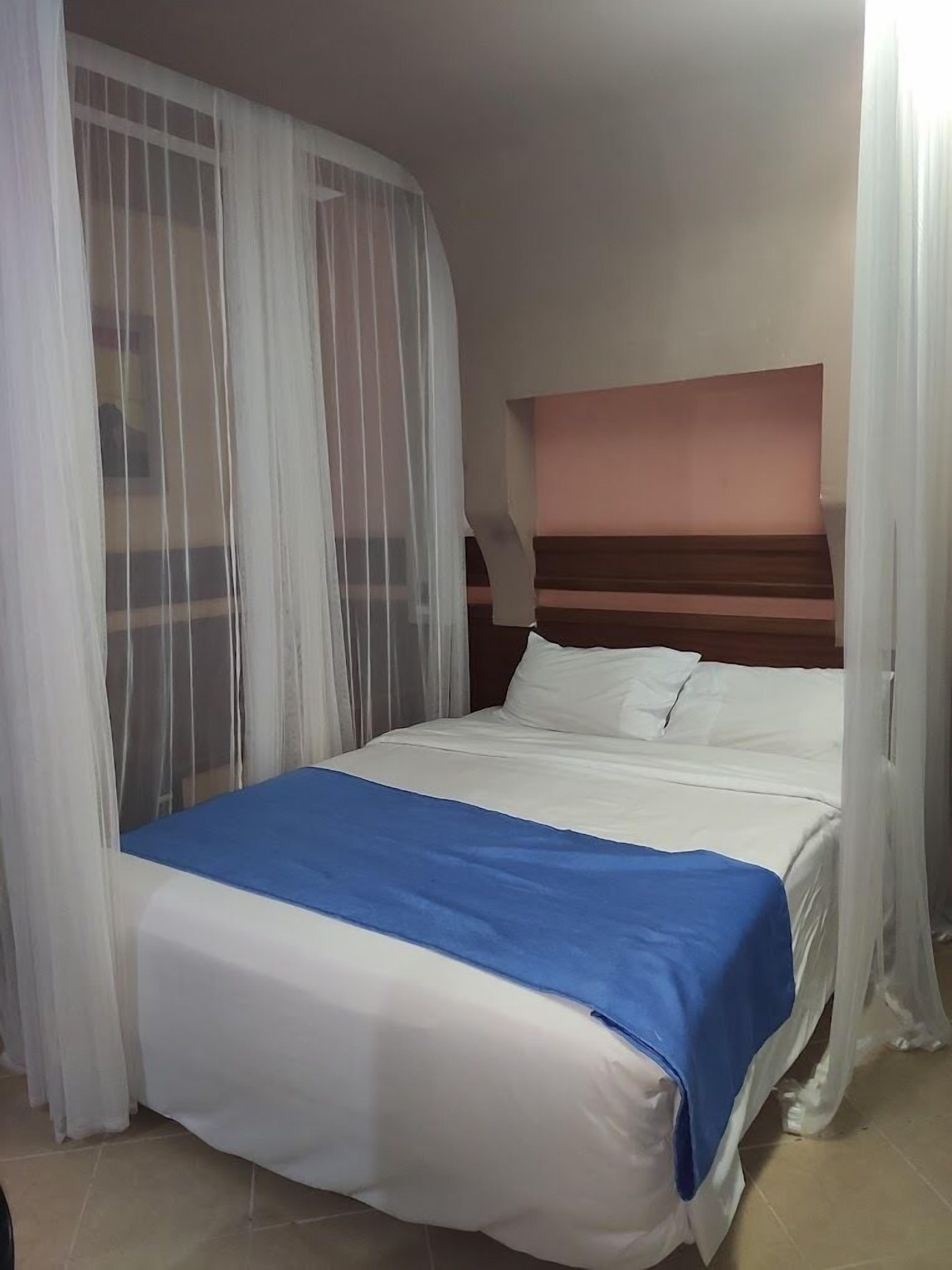 Een bed met wit- en blauwe beddengoed, een houten hoofdbord en lichte witte gordijnen.