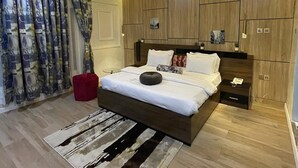 A modern bedroom with a large bed, a red ottoman, a wooden wardrobe, and a ceiling light fixture.