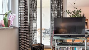 A living room with a TV on a glass table, a bookshelf with DVDs, a suitcase, and curtains. - Ammerside by Interhome (Weilheim in Oberbayern)
