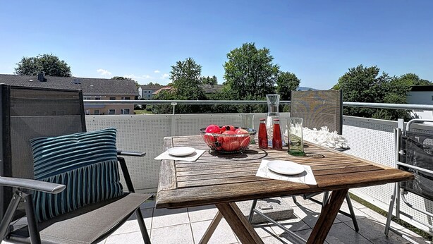 A wooden table with a white tablecloth, a bowl of fruit, and two bottles on a balcony with a view of houses and trees. - Ammerside by Interhome (Weilheim in Oberbayern)