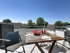 Una mesa de madera con mantel blanco, un cuenco de fruta y dos botellas en un balcón con vistas a casas y árboles.
