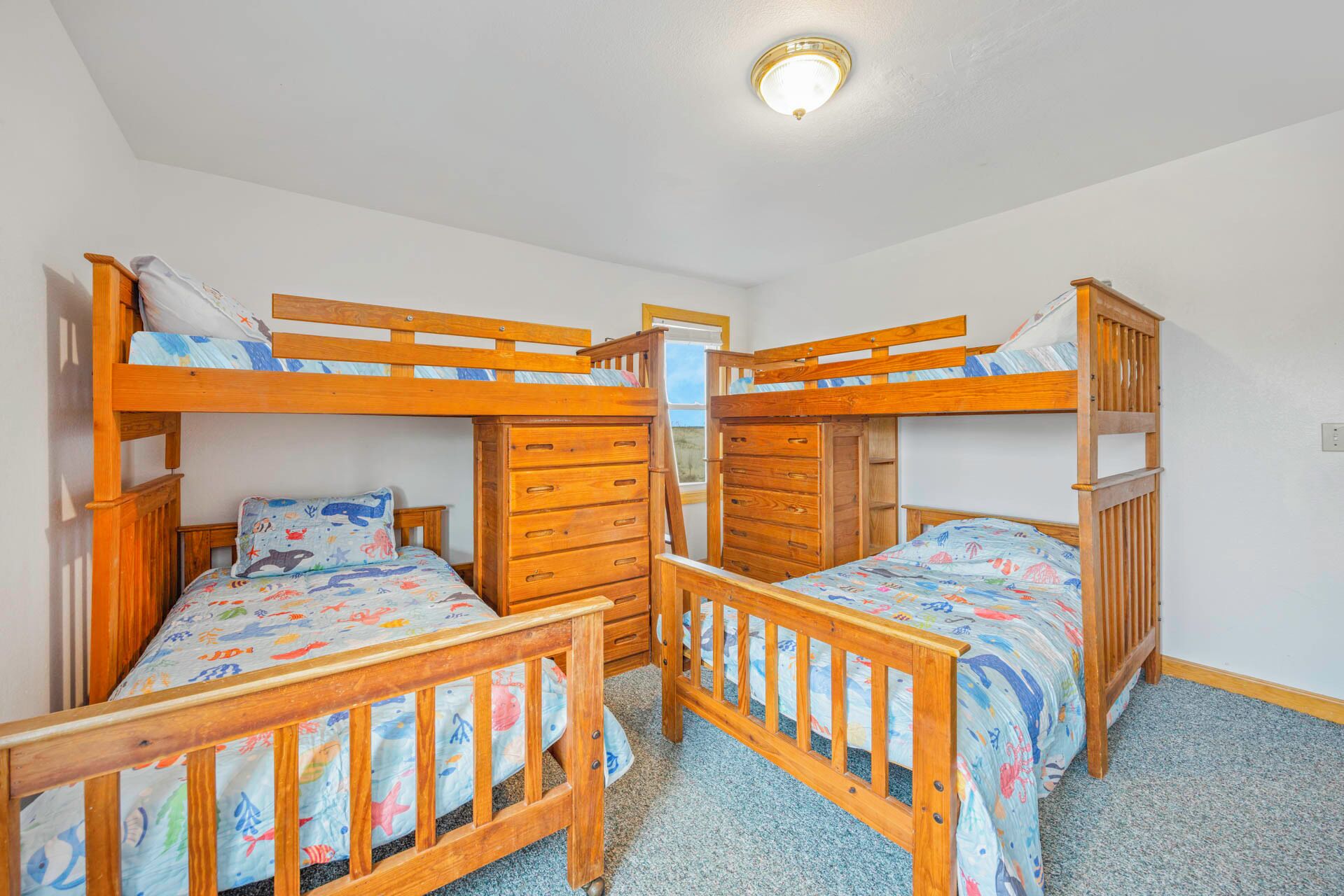 A room with three bunk beds, each with a wooden frame and a colorful bedspread. There is a wooden dresser with drawers and a mirror. The room has white walls and a ceiling light fixture.