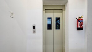 A narrow hallway with a door at the end, a fire extinguisher mounted on the wall, and a small light switch on the wall. - Super Hotel O Valasaravakkam Metro Mall (Chennai)