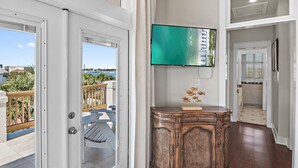 A room with a large TV mounted on the wall, a wooden cabinet with decorative items, and a view of the outdoors through a door. - White Sails | 3BR Coastal Retreat in Destin Pointe (Destin)
