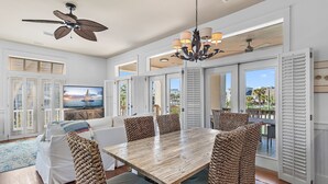 A spacious dining area with a wooden table and woven chairs, large windows with shutters, and a ceiling fan. - White Sails | 3BR Coastal Retreat in Destin Pointe (Destin)