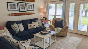Ein Wohnzimmer mit einem dunkelblauen Sofa, einem beigen Sessel und einem Couchtisch. An der Wand hängen gerahmte Bilder und ein Surfbrett ist zu sehen.