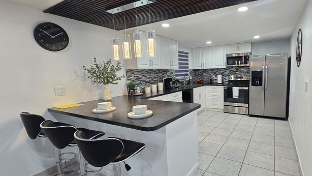 A modern kitchen with a central island, black bar stools, stainless steel appliances, and a clock on the wall. - #39 The Farmington House (Port Richey)