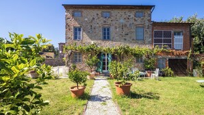 Un bâtiment en pierre avec une porte centrale, une extension en briques et un chemin de gravier menant à un espace aménagé pour s’asseoir.