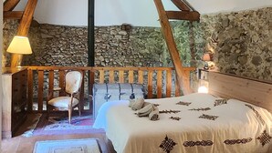 Ein rustikales Schlafzimmer mit sichtbaren Holzbalken, einer Steinwand, einem Bett mit gemustertem Überwurf, einem Sessel und einer Lampe.