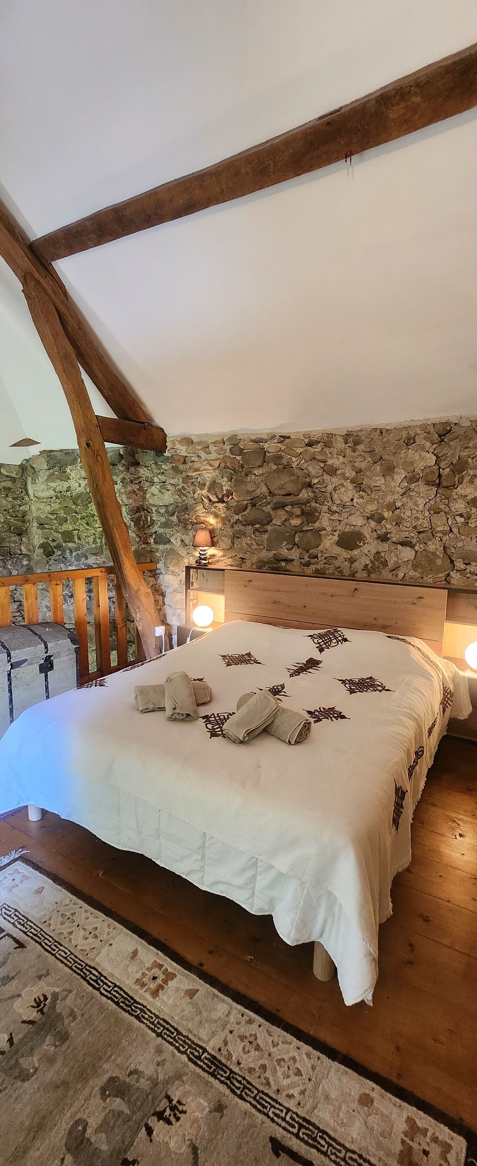 Une chambre avec un mur en pierre, des poutres apparentes, un lit avec une literie blanche et une table de chevet avec une lampe.