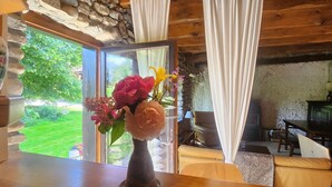 Ein rustikales Interieur mit einem Holztisch, einer Blumenvase und Blick auf einen Garten durch eine Glastür.