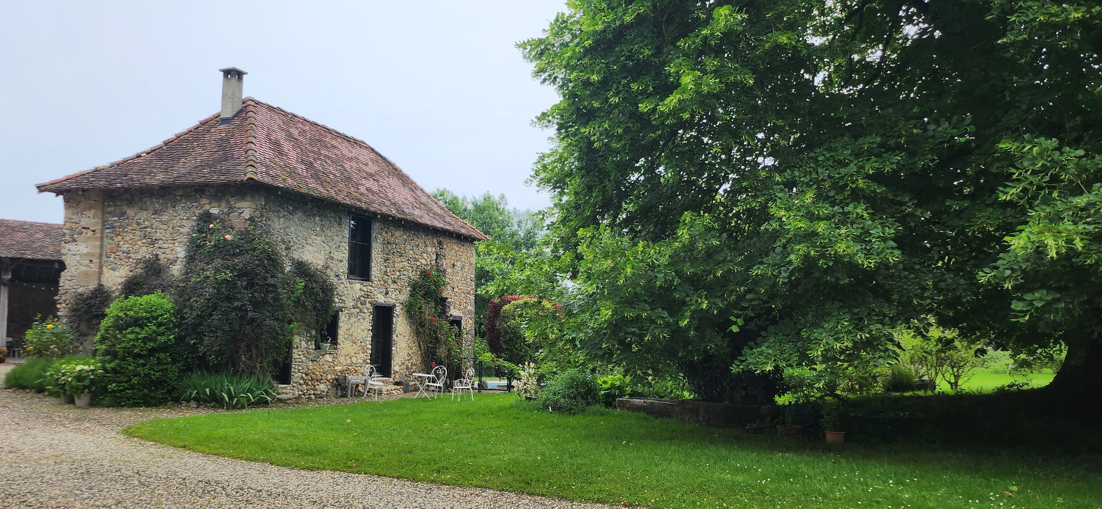 A stone building with a tiled roof, surrounded by a well-maintained garden and a large tree.