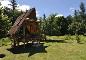 Une construction rustique en bois, dotée d’un toit de chaume, entourée de verdure et d’arbres.