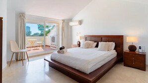 Une chambre spacieuse avec un grand lit, un coin salon avec un fauteuil et une table, et un balcon agrémenté de rideaux.