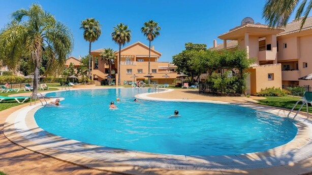 Une piscine entourée de chaises longues et de palmiers, le tout bordé par un bâtiment d’hôtel.
