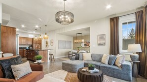 A spacious living area with a grey sofa, a brown armchair, and a coffee table. There is a kitchen in the background with wooden cabinets and a dining area with a table and chairs. - Ember & Ash Designer Home w/ King Beds, Games (Fort Collins)