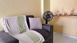 A sofa with a blanket, a standing fan, and a shelf with glasses and cups. - AirBNBusiness Maracanaú (Maracanau)