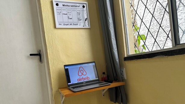 A laptop on a wooden shelf with the Airbnb logo, a calendar on the wall, and a window with a metal grid.