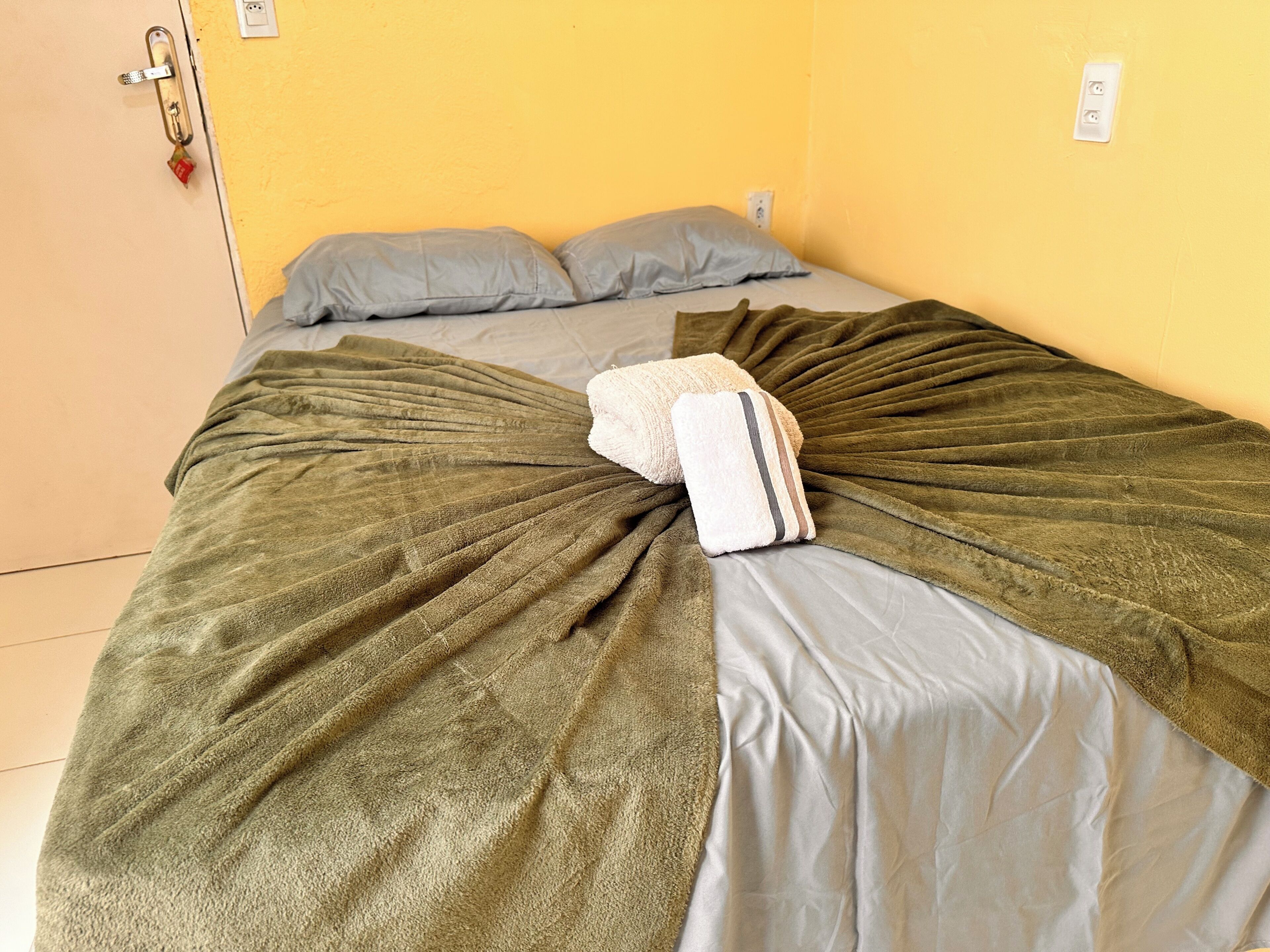 A bed with a green blanket and pillows, a door with a lock, and a wall socket.