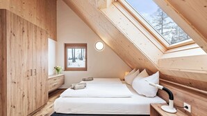 Un dormitorio con techo inclinado, tragaluces, cama, mesitas de noche y ventana con vistas a árboles.