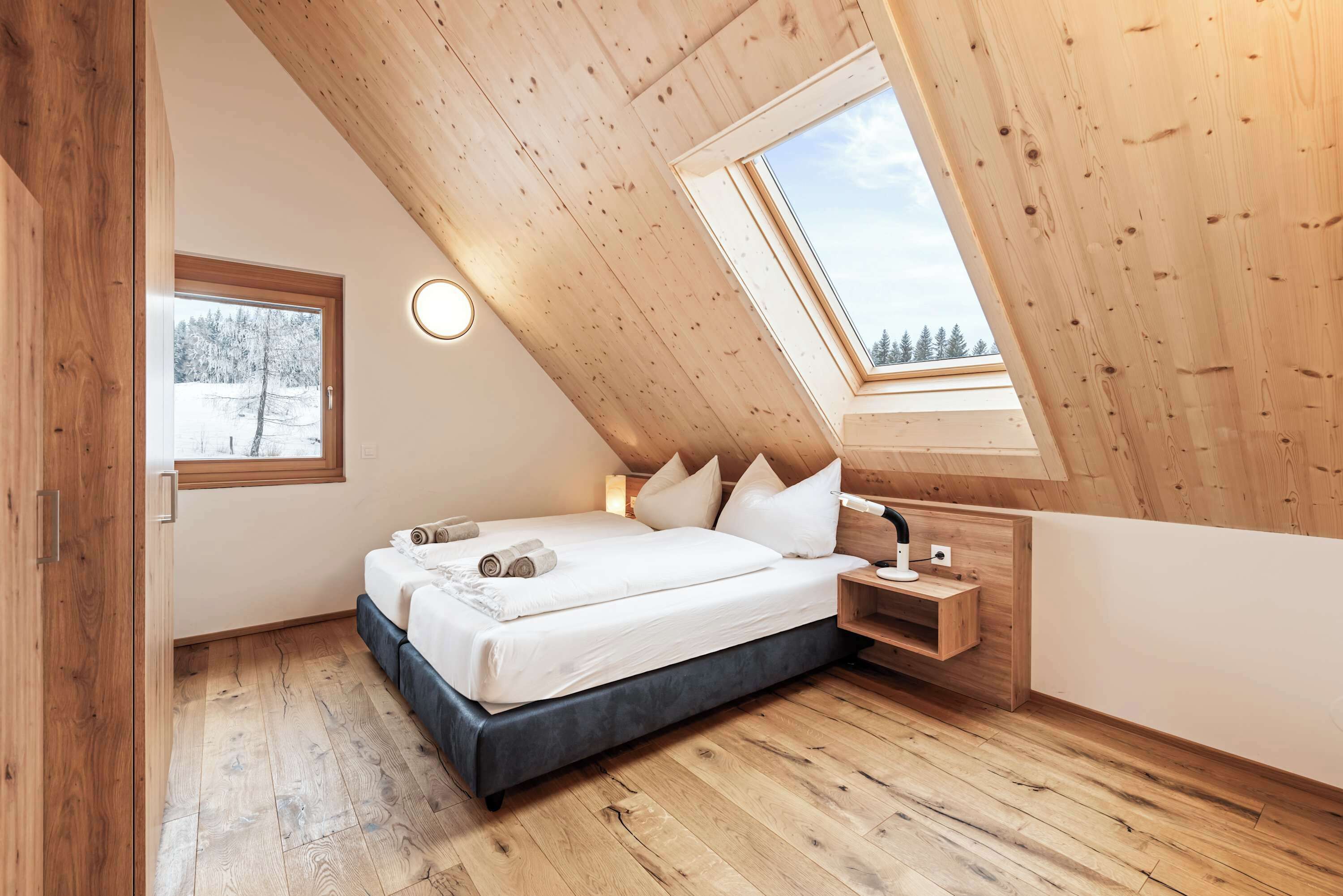 Un dormitorio con techo inclinado, tragaluz, cama con almohadas, mesita de noche y ventana con vista a árboles.