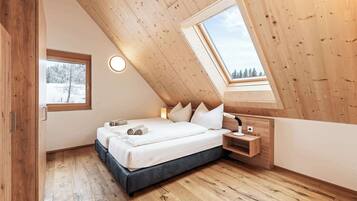 Un dormitorio con techo inclinado, tragaluz, cama con almohadas, mesita de noche y ventana con vista a árboles.