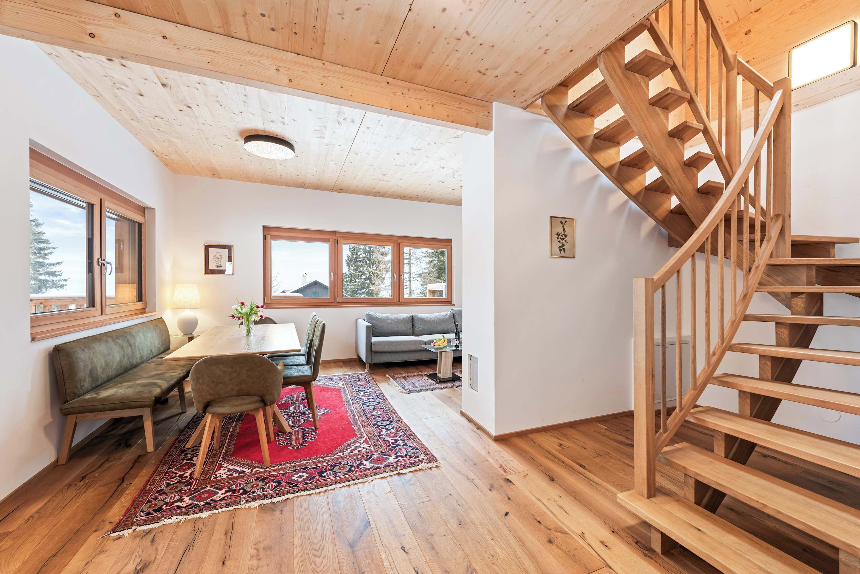 A spacious living area with a wooden staircase, a sofa, a dining table with chairs, and large windows.